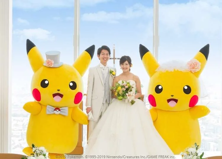 Couple japonais en tenue de mariage entouré de deux Pikachu, un en costume de marié avec haut-de-forme et l'autre en robe de mariée avec voile