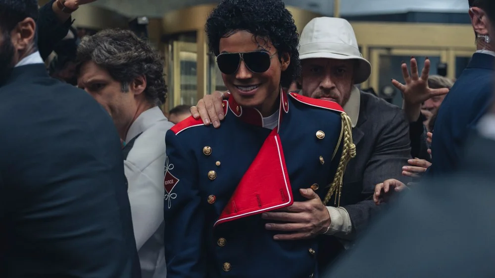 Jaafar Jackson en Michael Jackson, veste militaire bleue et lunettes aviateur, entouré de gardes du corps dans une scène du film