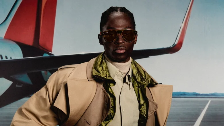 Un homme en trench-coat porte des Ray-Ban Meta Gen 3 à monture aviateur devant un avion de ligne
